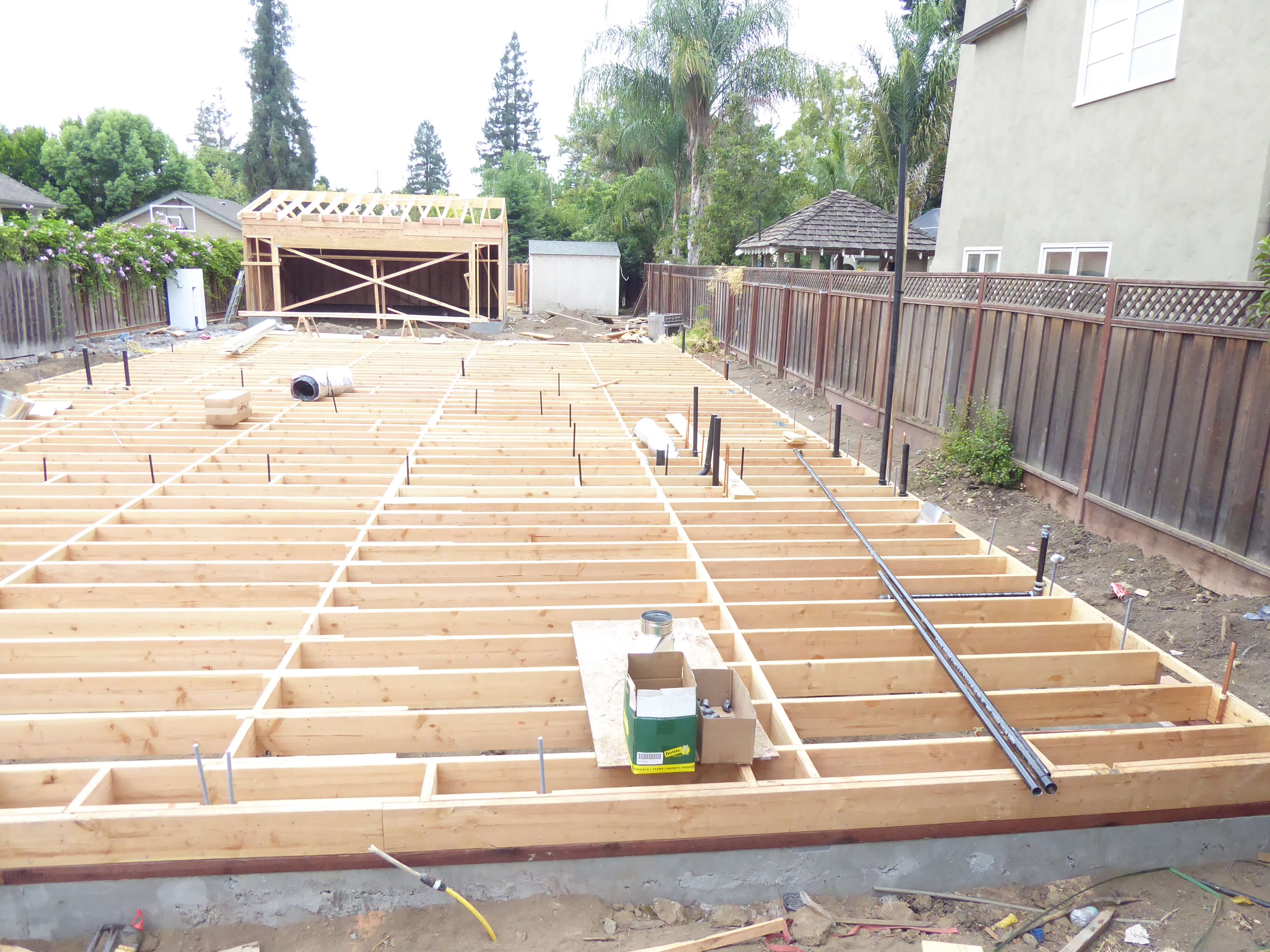Home construction site with raised wood floor framing, plumbing rough-ins, and detached structure under renovation in backyard.